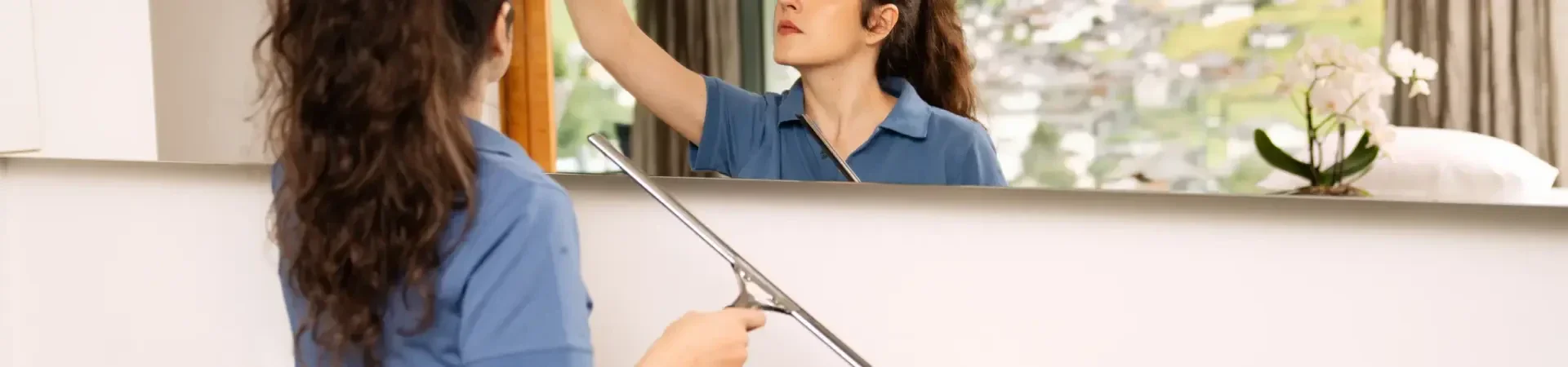 Eine Person in einer blauen Uniform reinigt einen großen Wandspiegel mit einem Abzieher.
