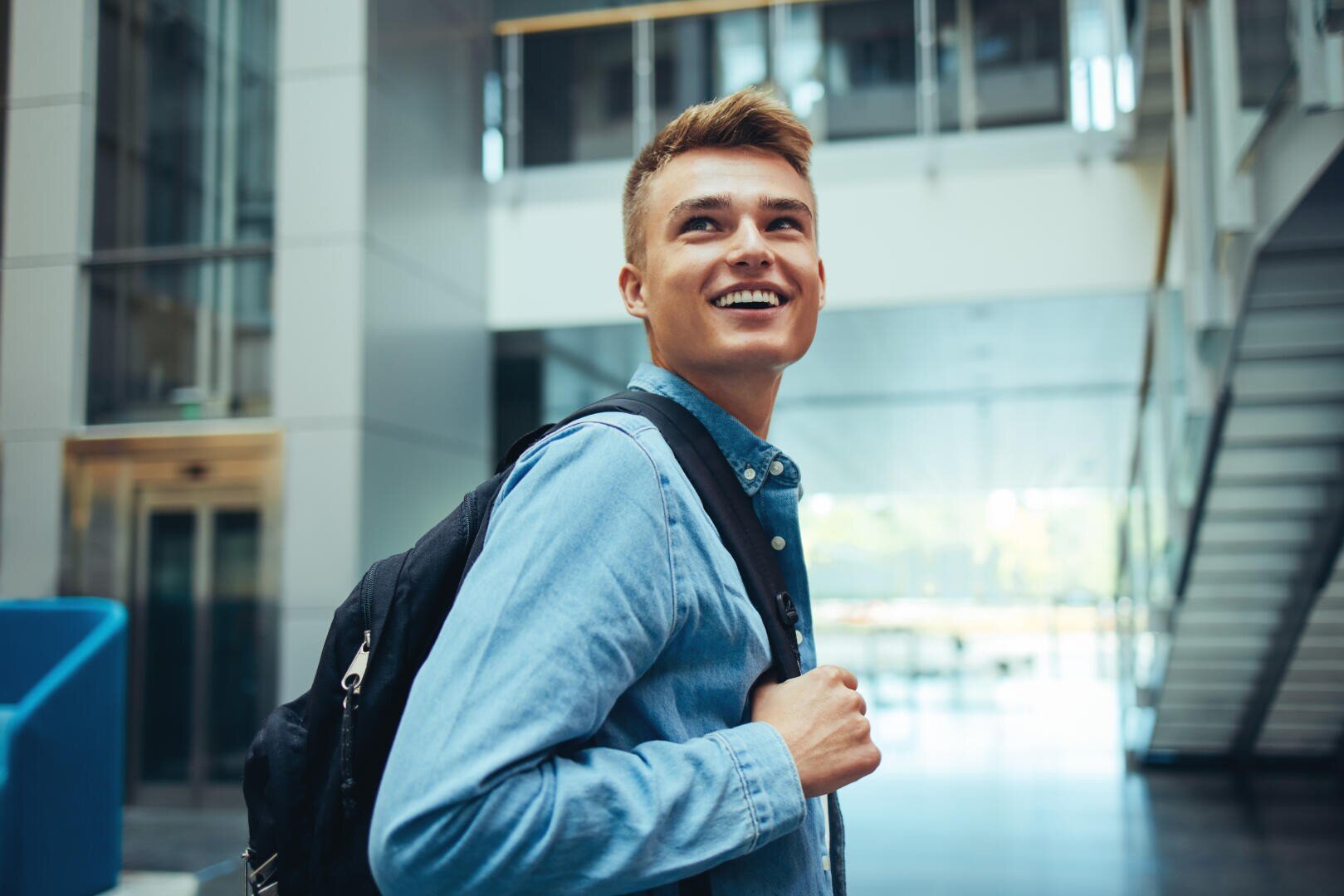 Ein junger Mann mit kurzen Haaren, der ein Jeanshemd trägt und einen Rucksack mit sich führt, lächelt, während er in einem modernen, hellen Gebäude mit Glaswänden und einem Aufzug im Hintergrund steht.