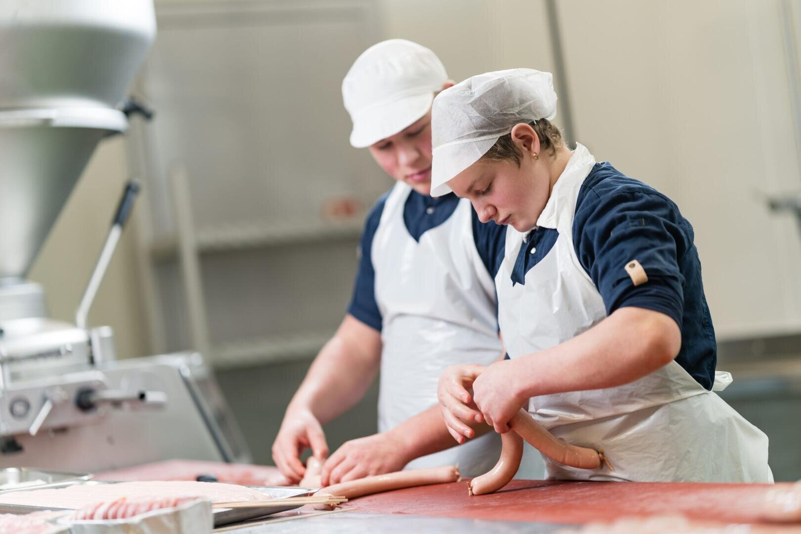 In einer Produktionsstätte arbeiten zwei Personen, die weiße Schürzen und Haarnetze tragen, an einem Tisch und formen Würste.