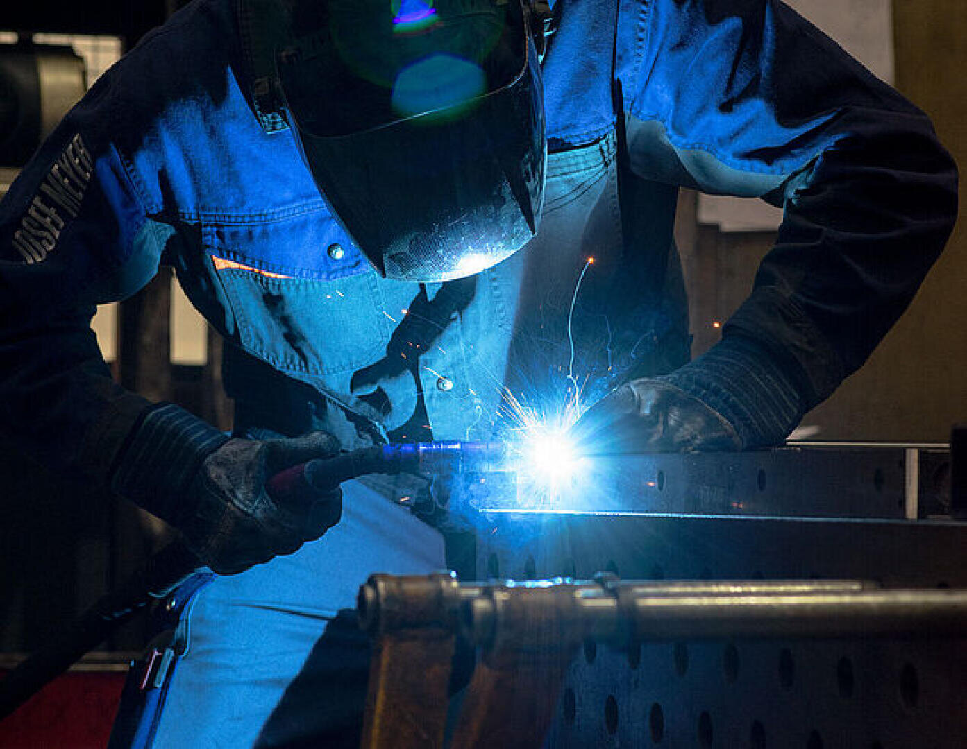 Eine Person, die Schutzkleidung und einen Schweißhelm trägt, bedient einen Schweißbrenner und erzeugt dabei hellblaue Funken und Licht, während sie in einer industriellen Umgebung an einer Metalloberfläche arbeitet.