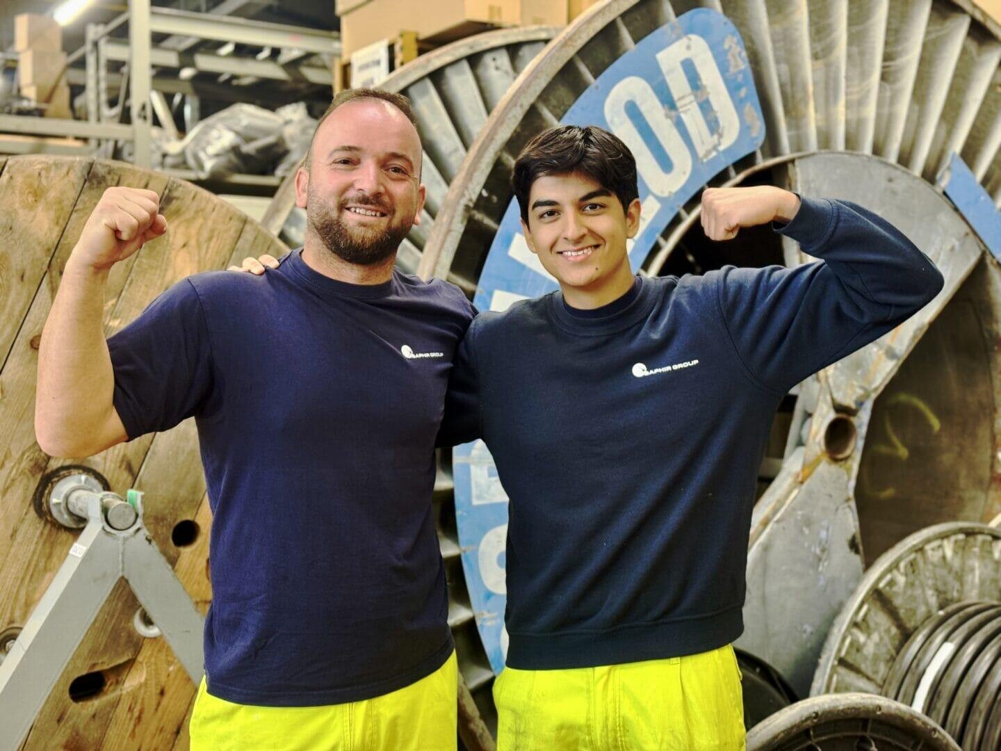 Zwei Männer in dunkelblauen Hemden und gelben Hosen stehen nebeneinander in einer Industrieumgebung, lächeln und spannen ihre Arme an. Im Hintergrund sind große Spulen und Maschinen zu sehen.