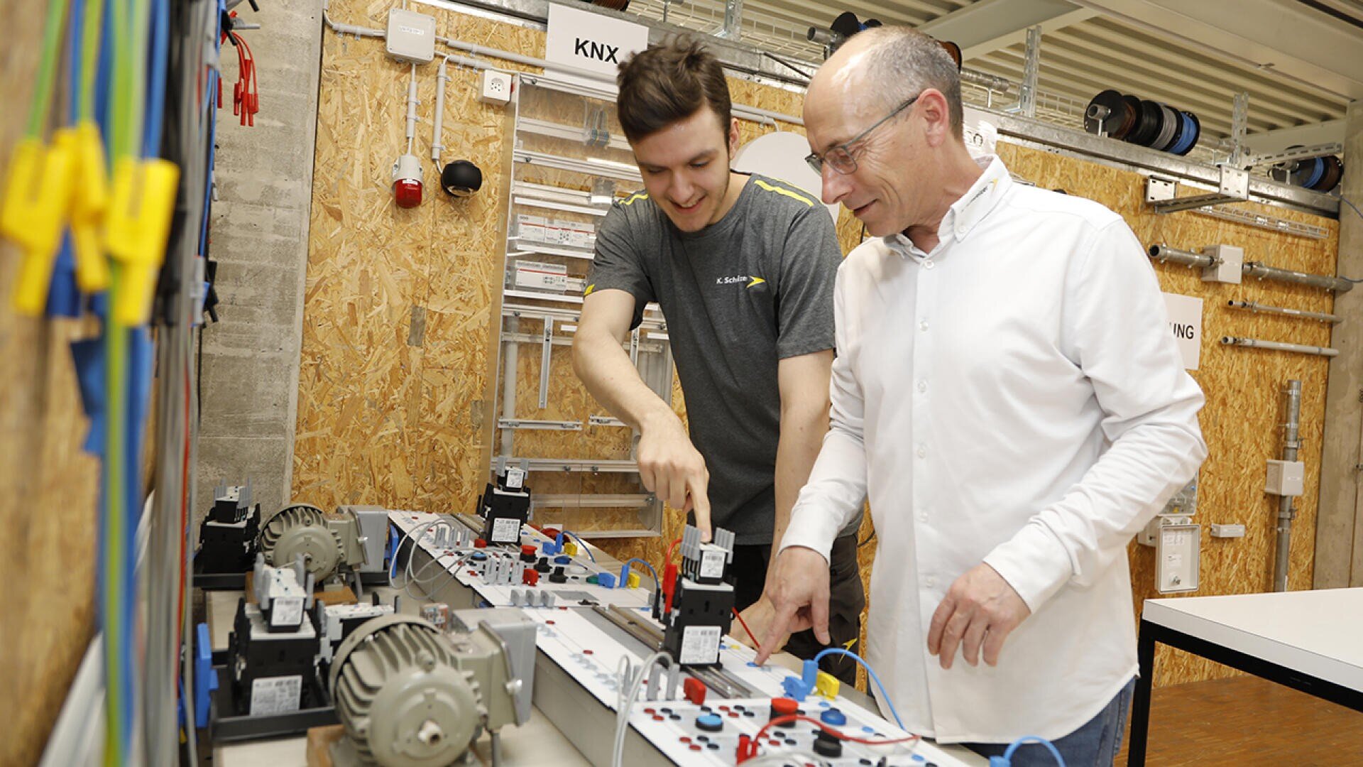 Zwei Männer arbeiten gemeinsam an einem elektrotechnischen Projekt, indem sie in einer Werkstattumgebung mit sichtbaren Werkzeugen und Geräten Komponenten an einer Schalttafel einstellen.
