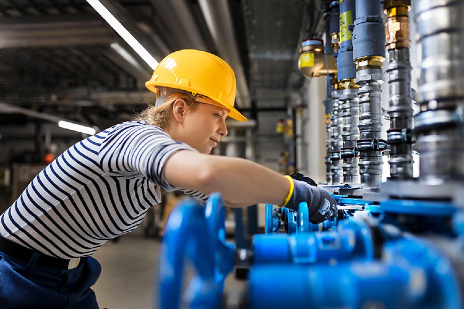 Eine Person mit einem gelben Schutzhelm und einem gestreiften Hemd arbeitet an Industriemaschinen und justiert Ventile und Steuerungen in einer Fabrik oder einem Werk.
