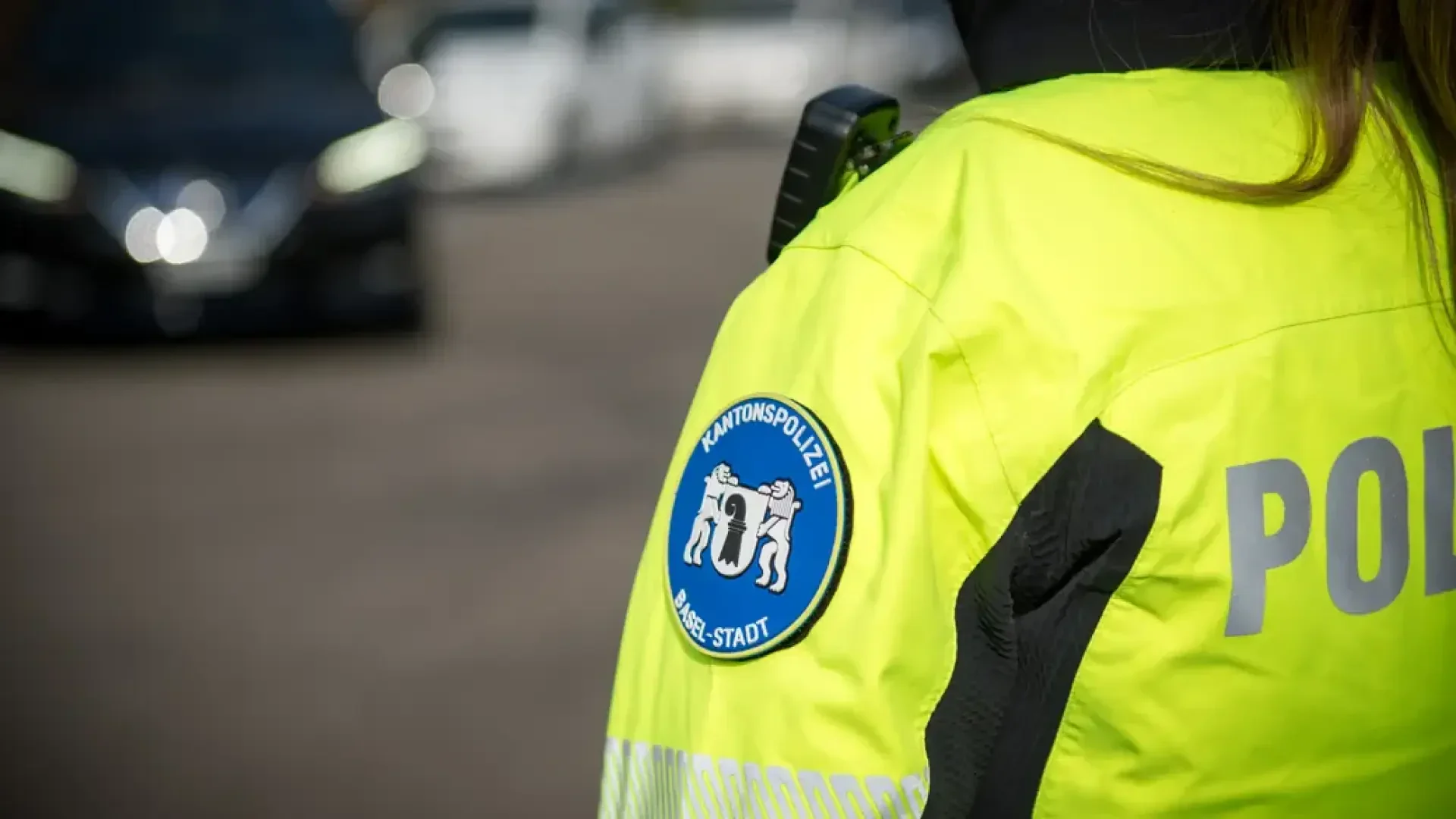 Eine Nahaufnahme eines Polizeibeamten, der eine leuchtend gelbe Jacke mit einem blauen Aufnäher am Ärmel trägt und vor einer unscharfen Straße mit Autos im Hintergrund steht.