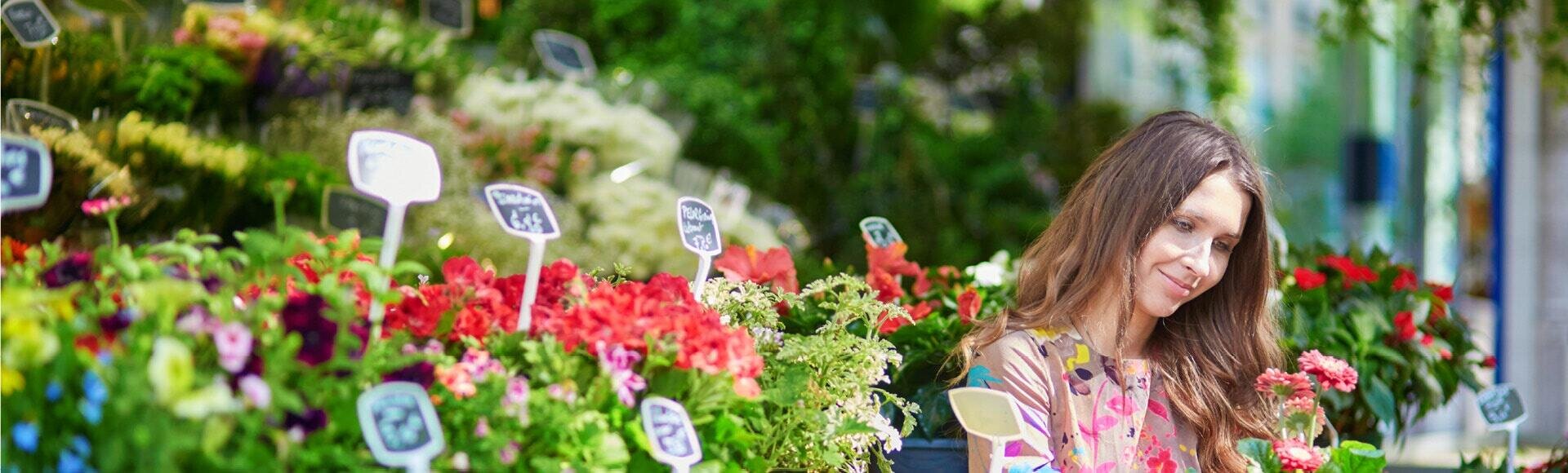 Eine Frau in einem geblümten Hemd lächelt, während sie auf einem Markt unter freiem Himmel Topfpflanzen betrachtet, umgeben von bunten Blüten und Pflanzenschildern.