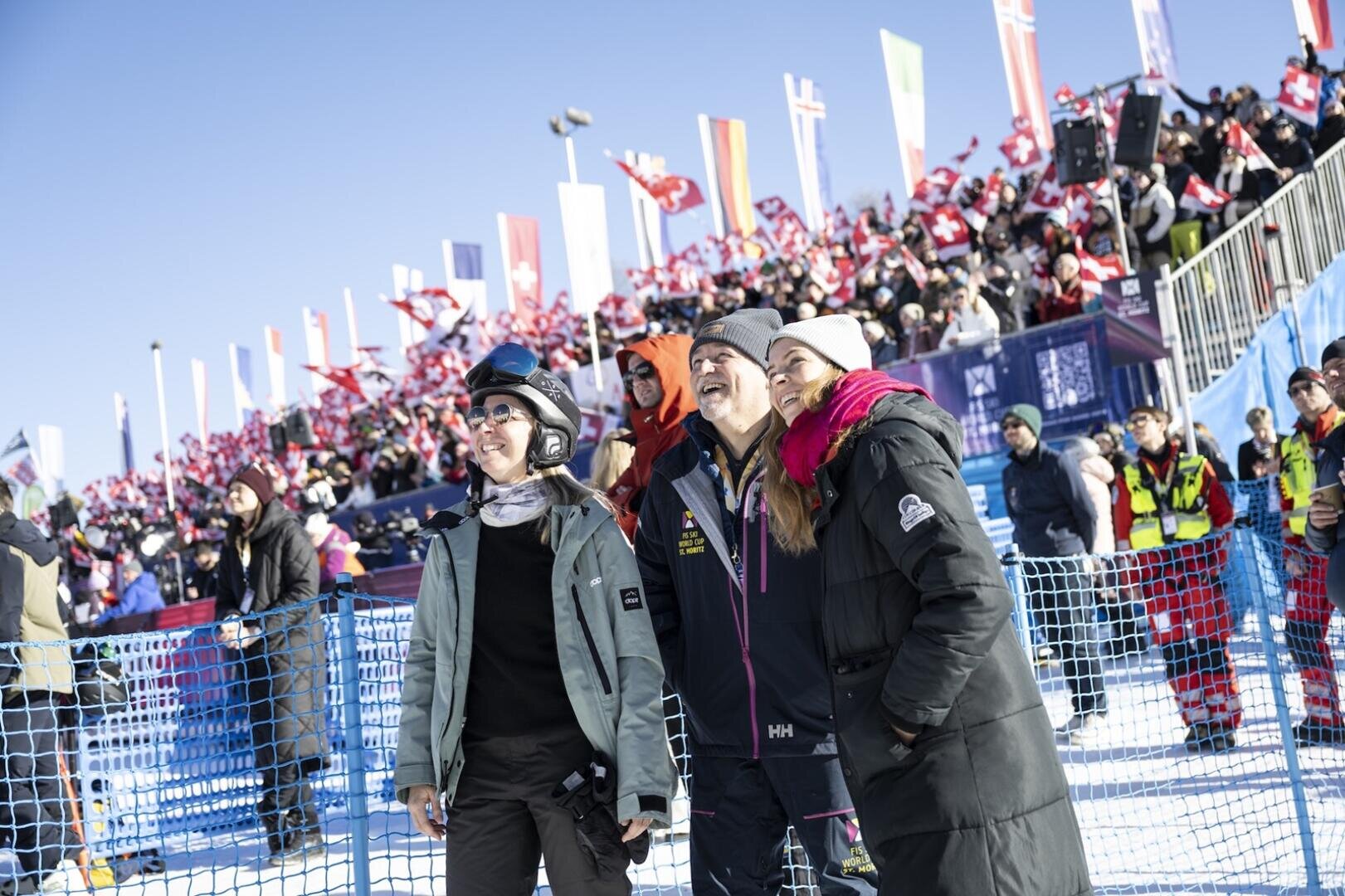 Drei Menschen in Winterkleidung stehen lächelnd vor einer jubelnden Menschenmenge mit Fahnen am FIS Skiweltcup St. Moritz.