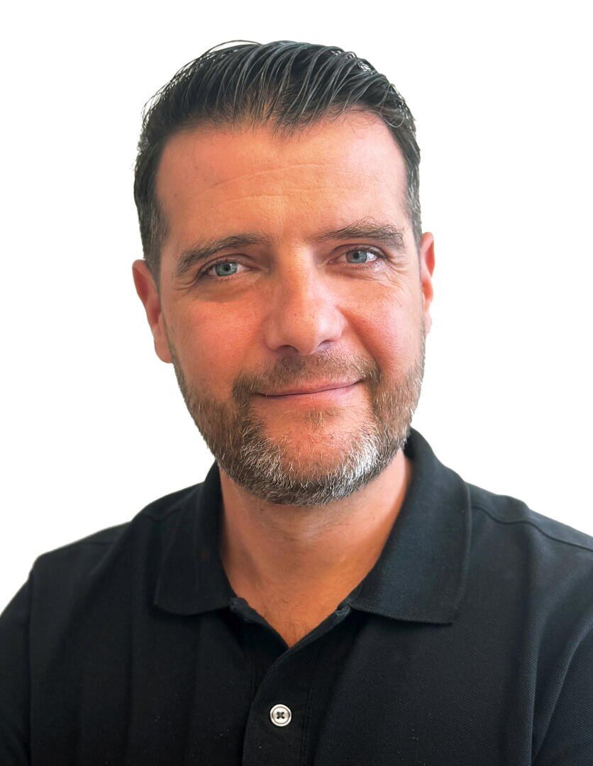 Ein Mann mit dunklem Haar und einem hellen Bart, der lächelt und ein schwarzes Poloshirt trägt.
