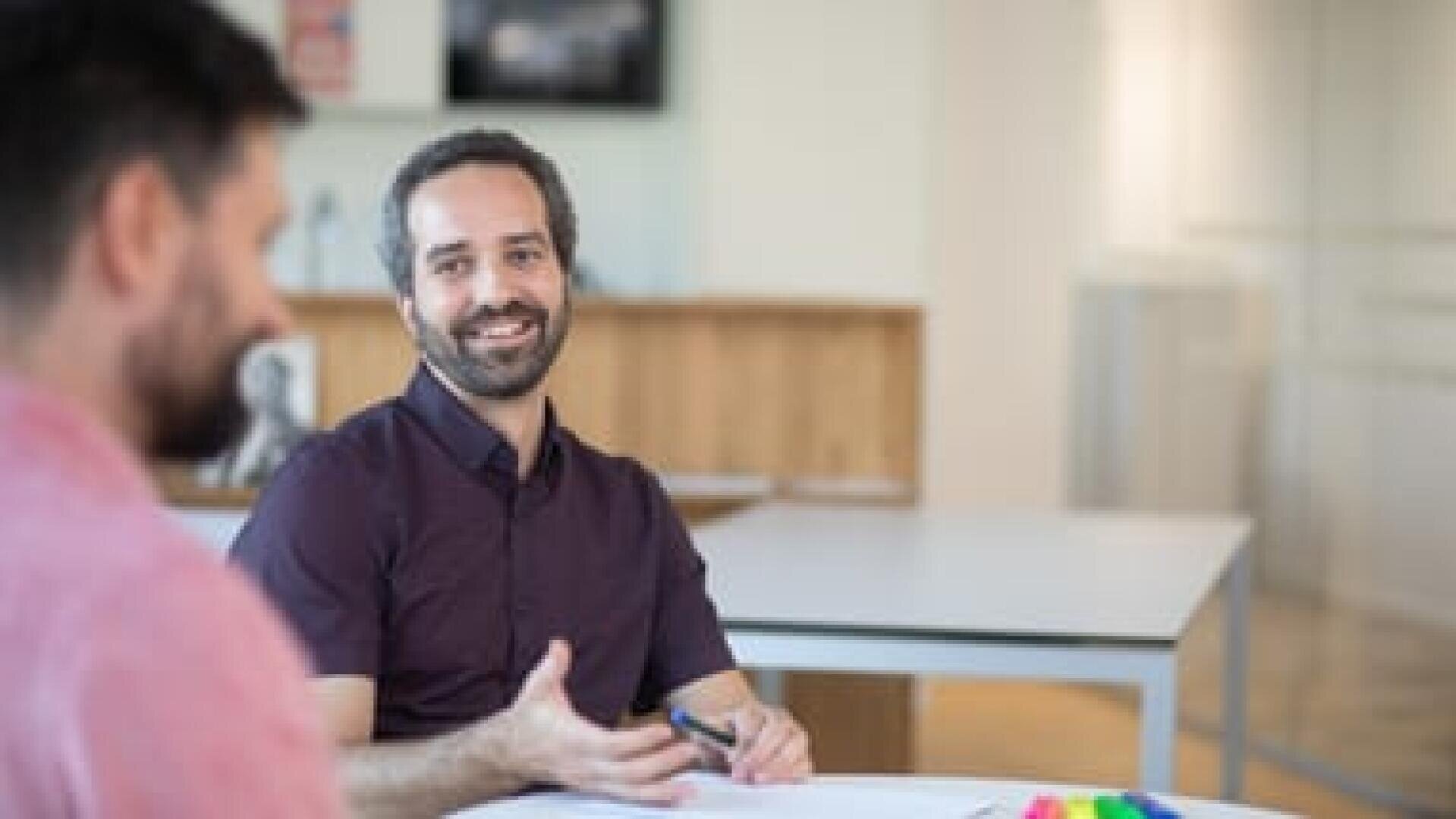 Ein Mann mit kurzen dunklen Haaren und Bart, der ein dunkles Hemd trägt, lächelt und gestikuliert, während er sich mit einem anderen Mann in einem hellen Raum unterhält, in dessen Hintergrund Tische und Regale zu sehen sind.