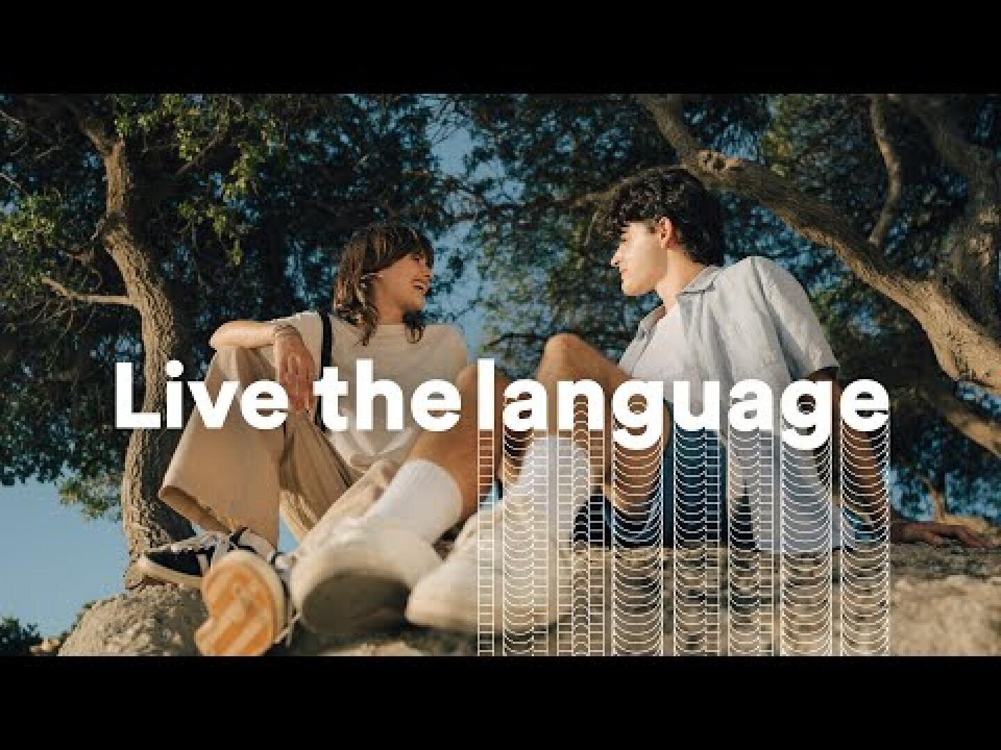 Zwei junge Menschen sitzen auf Felsen unter Bäumen, lächeln und blicken einander an. Über dem Bild steht in weißer Schrift der Text „Live the language“, unter den Worten sind dekorative Wellenlinien zu sehen.