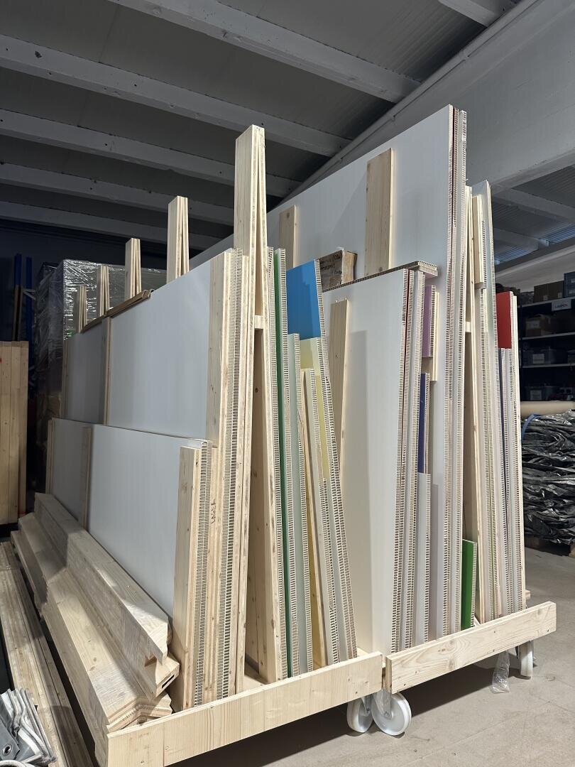 Große durchsichtige und undurchsichtige Kunststoffplatten verschiedener Größen werden aufrecht in einem Holzrahmen auf Rädern in einem Industrielager gelagert, wobei im Hintergrund Regale und Materialien zu sehen sind.