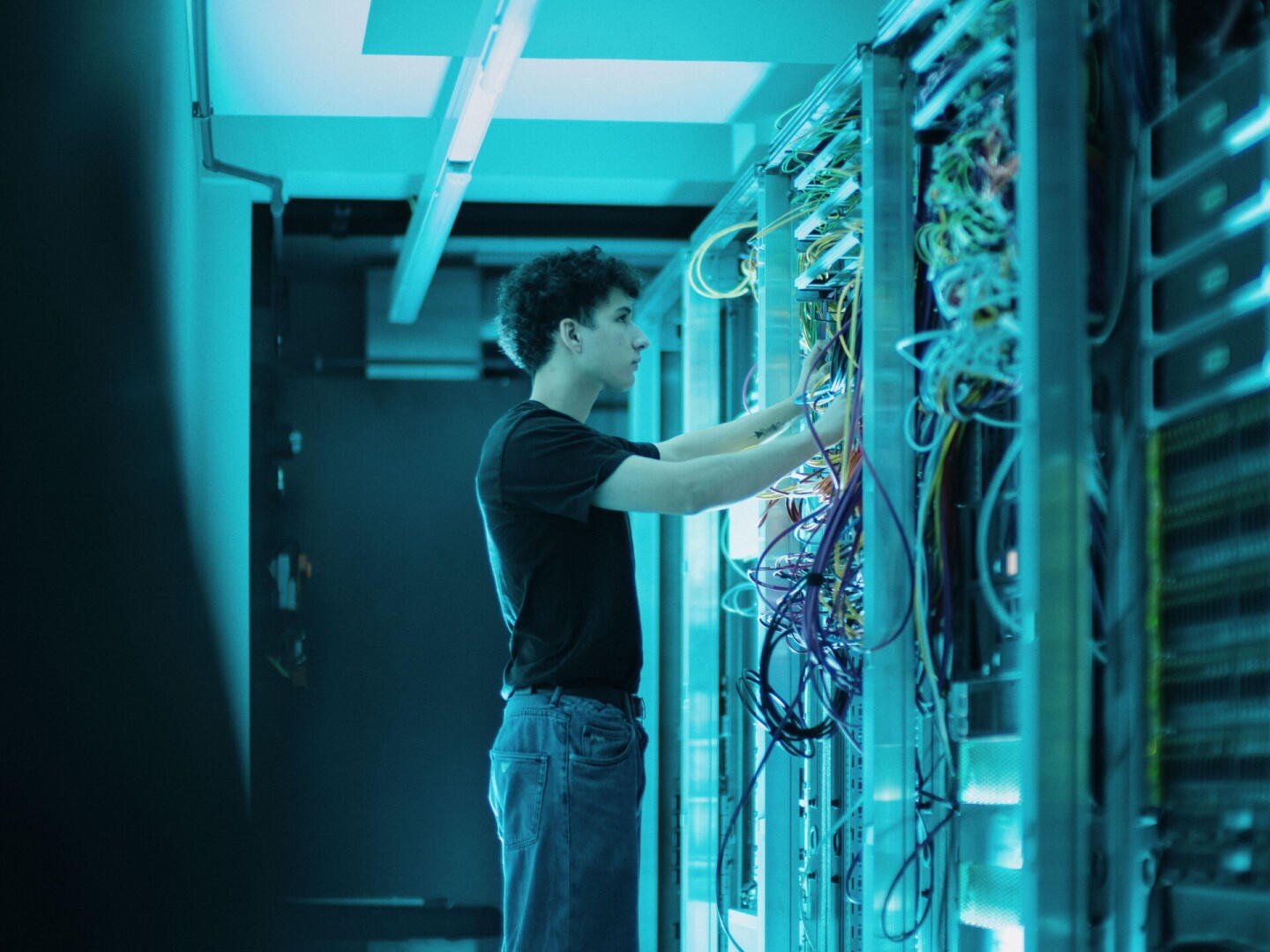 Eine Person in einem schwarzen Hemd arbeitet an einem Serverrack in einem schwach beleuchteten Raum und schließt Netzwerkkabel an verschiedene elektronische Geräte an oder ordnet sie.