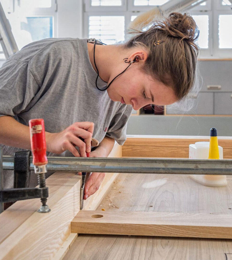 Eine Person bearbeitet in einer hellen Werkstatt mit einem Handwerkzeug und einer Klemme einen Holzrahmen. Holzspäne sind zu sehen, und die Person scheint ganz auf ihre Arbeit konzentriert zu sein.