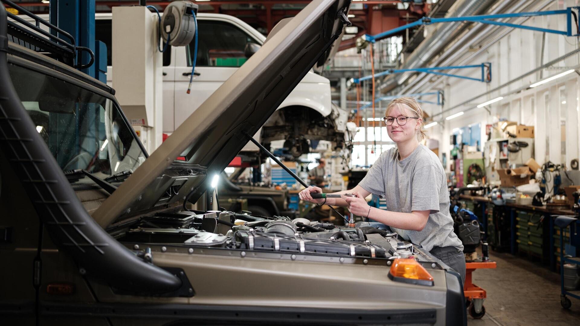 Eine Person mit Brille und in einem grauen Hemd arbeitet in einer geschäftigen Autowerkstatt an einem Automotor, umgeben von Werkzeugen und anderen Fahrzeugen.