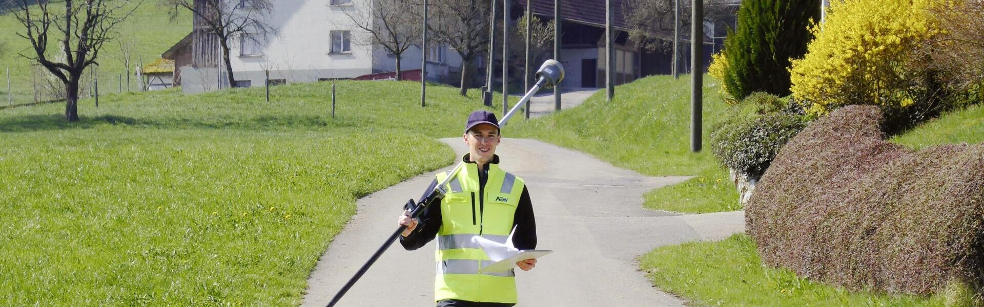 Eine Person in einer Warnweste und einer Mütze geht auf einem Feldweg entlang und hält Vermessungsgeräte sowie einen Schreibblock in der Hand; im Hintergrund sind grüne Felder und Gebäude zu sehen.