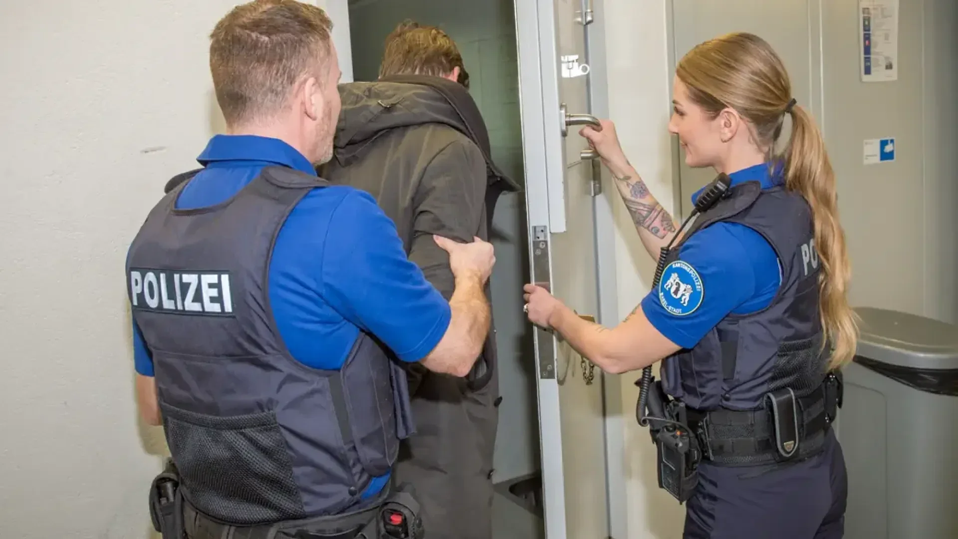 Zwei Polizeibeamte begleiten eine Person durch eine Tür. Die Beamten tragen blaue Uniformen mit der Aufschrift „Polizei“ und scheinen die Person, die mit dem Rücken zur Kamera steht, in einen gesicherten Bereich zu führen.