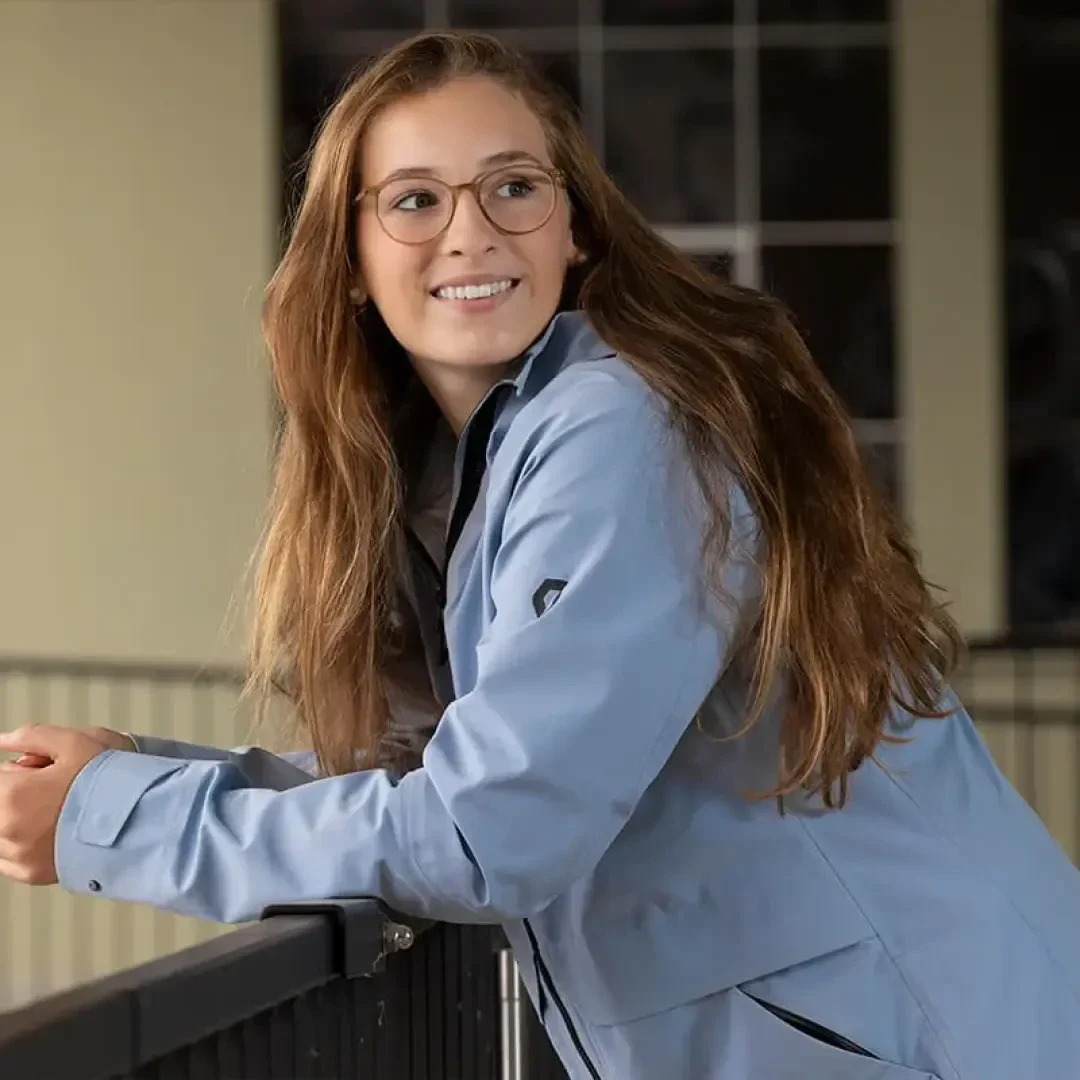 Eine Person mit langen braunen Haaren und Brille, die eine hellblaue Jacke trägt, lehnt an einem Geländer und lächelt, im Hintergrund ist ein verschwommenes Gebäude zu sehen.