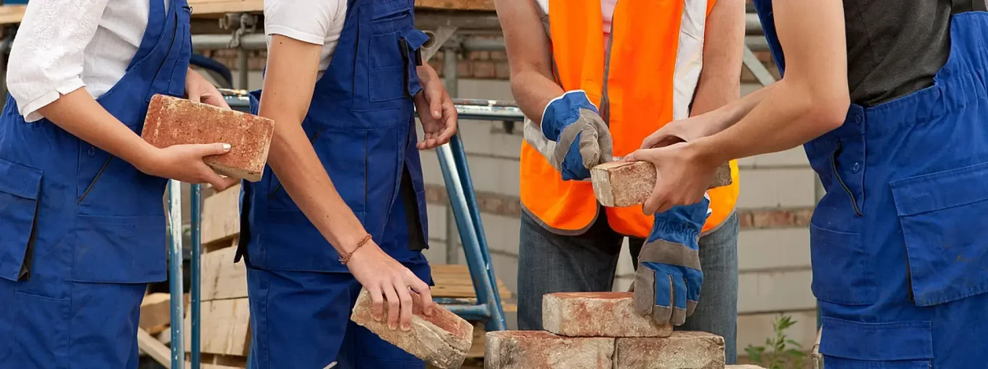 Vier Personen in blauen Overalls arbeiten gemeinsam daran, auf einer Baustelle rote Ziegelsteine zu stapeln.