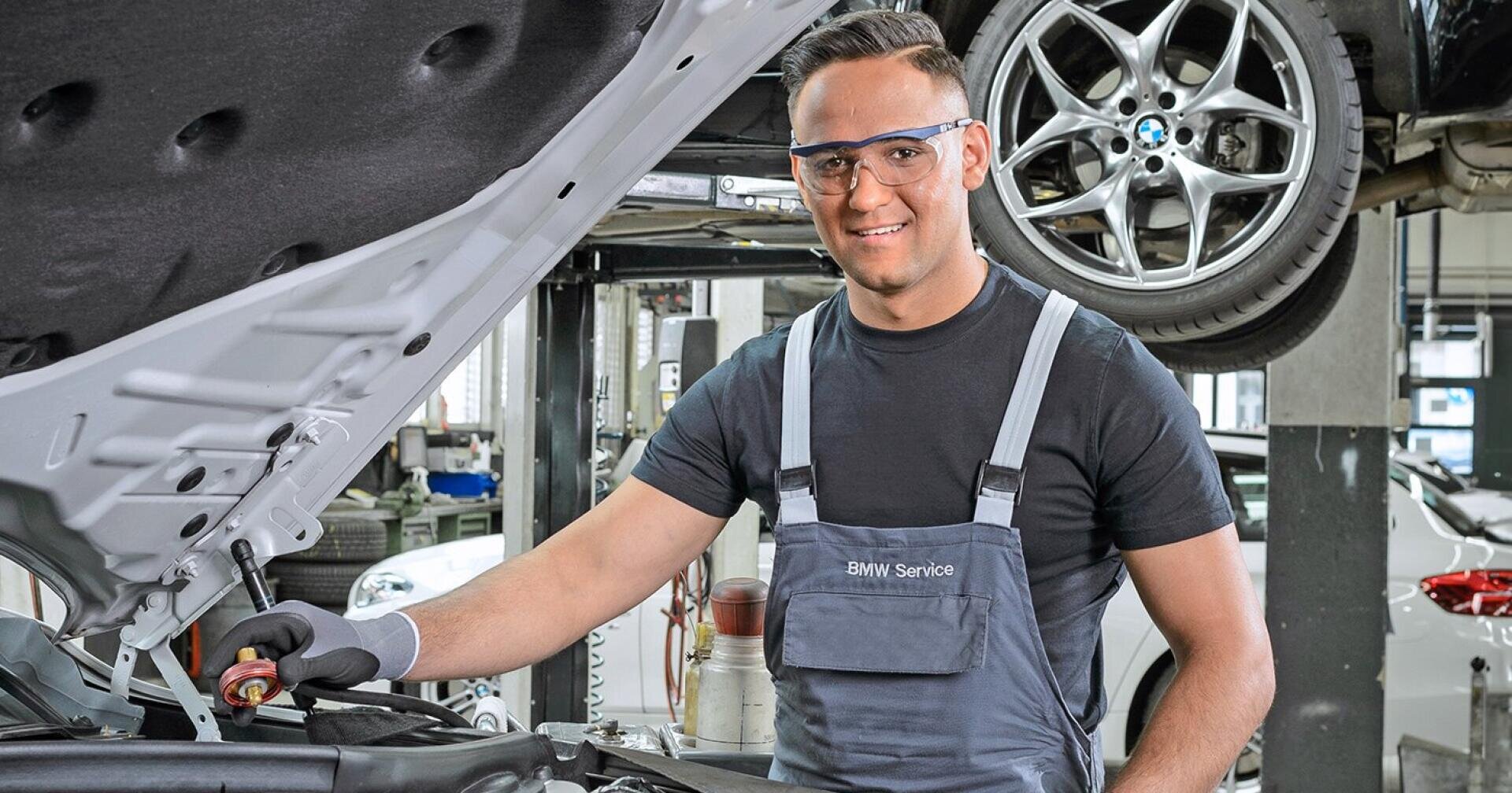 Ein Mechaniker, der Handschuhe und eine Schutzbrille trägt, lächelt, während er in einer BMW-Werkstatt an einem Auto arbeitet; im Hintergrund steht ein weiteres Auto auf der Hebebühne.