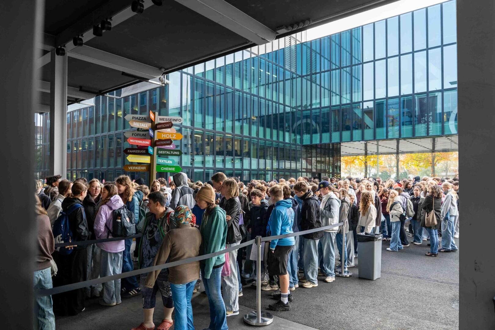 Eine große Menschenmenge, die draußen in einer langen Schlange vor einem modernen Glasgebäude neben einem Wegweiser mit verschiedenen Richtungsangaben steht.