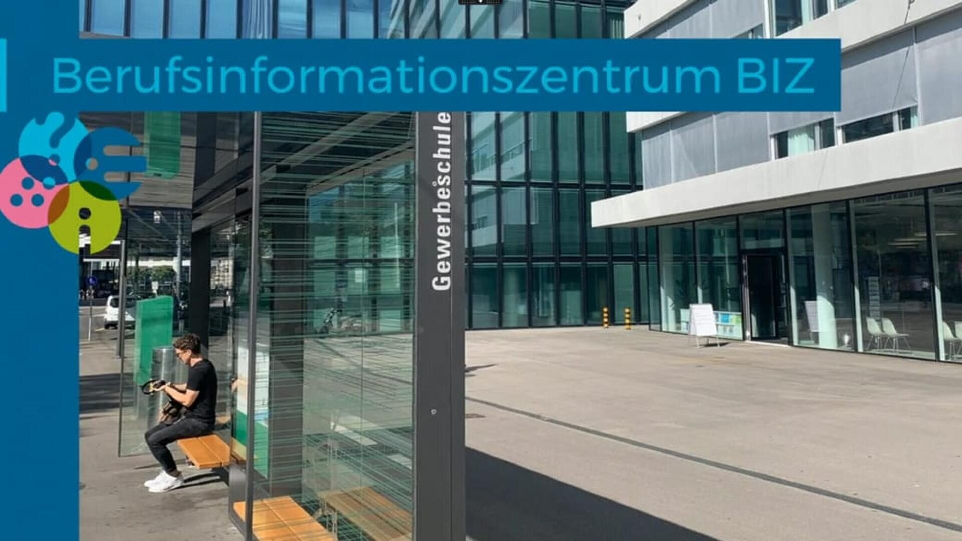 Ein Mann sitzt auf einer Bank an einer verglasten Bushaltestelle vor einem modernen Gebäude mit der Aufschrift „Gewerbeschule“. Auf einem blauen Banner steht „Berufsinformationszentrum BIZ“, links davon sind bunte Symbole zu sehen.