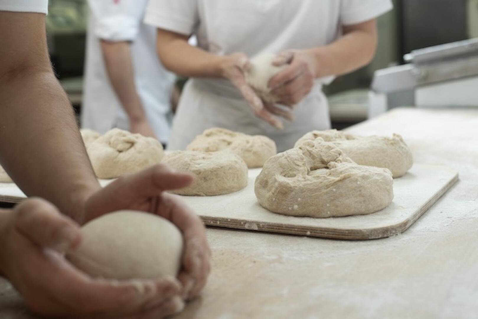Auf einem bemehlten Tisch kneten und formen Menschen Teigkugeln und bereiten in einer Bäckerei mehrere Brote zu.