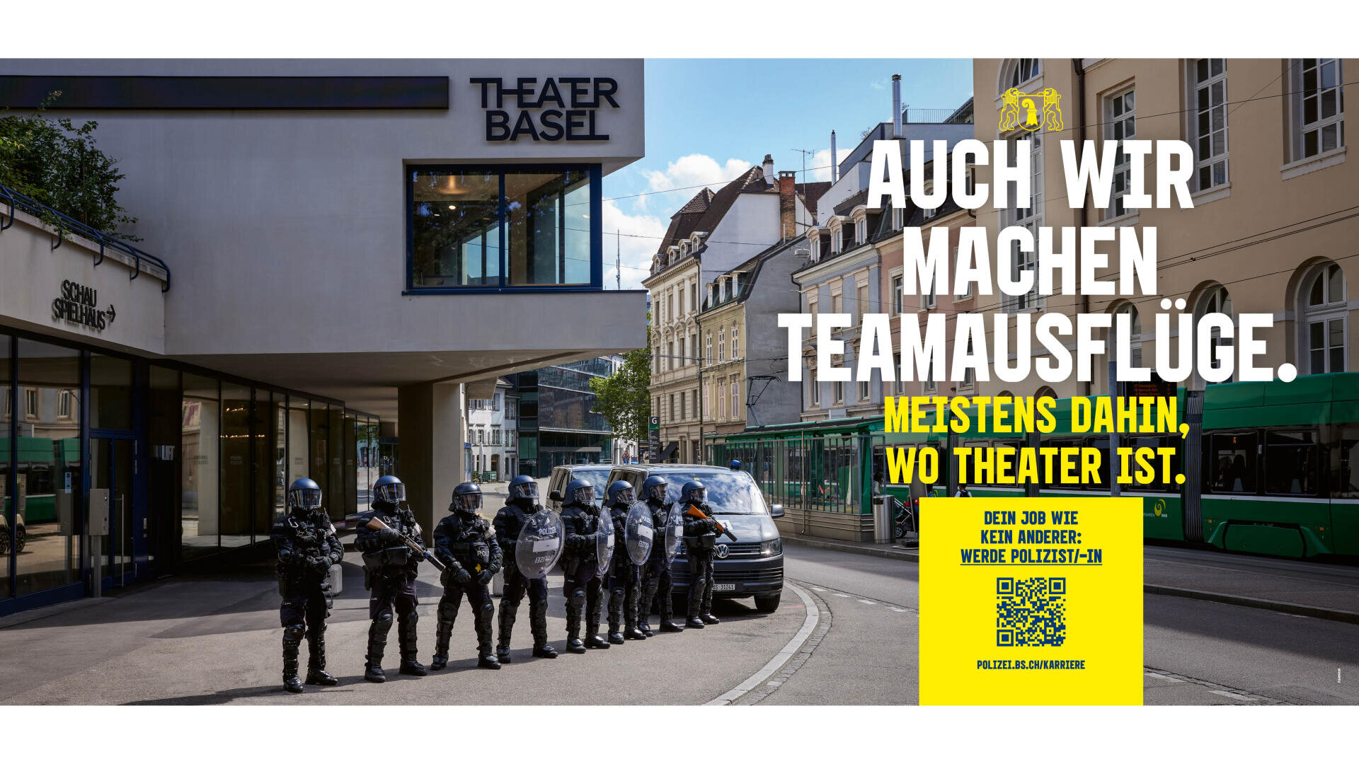 Vor dem Theater Basel steht eine Gruppe von Polizisten in Kampfausrüstung. Ein großer Schriftzug auf Deutsch lautet: „Auch wir machen Teamausflüge. Meistens dorthin, wo Theater ist.“ In der rechten unteren Ecke ist eine gelbe Stellenanzeige zu sehen.