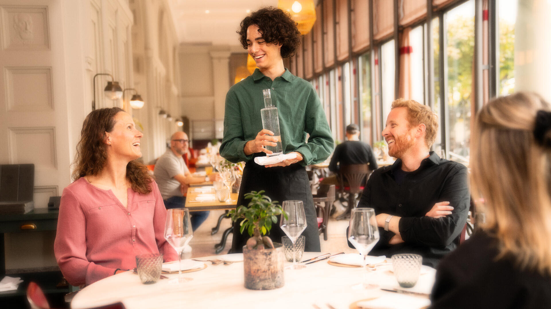 Ein lächelnder Kellner mit einer Flasche Wasser steht an einem Tisch mit drei fröhlichen Gästen in einem hellen, eleganten Restaurant mit großen Fenstern und ordentlich gedeckten Tischen.